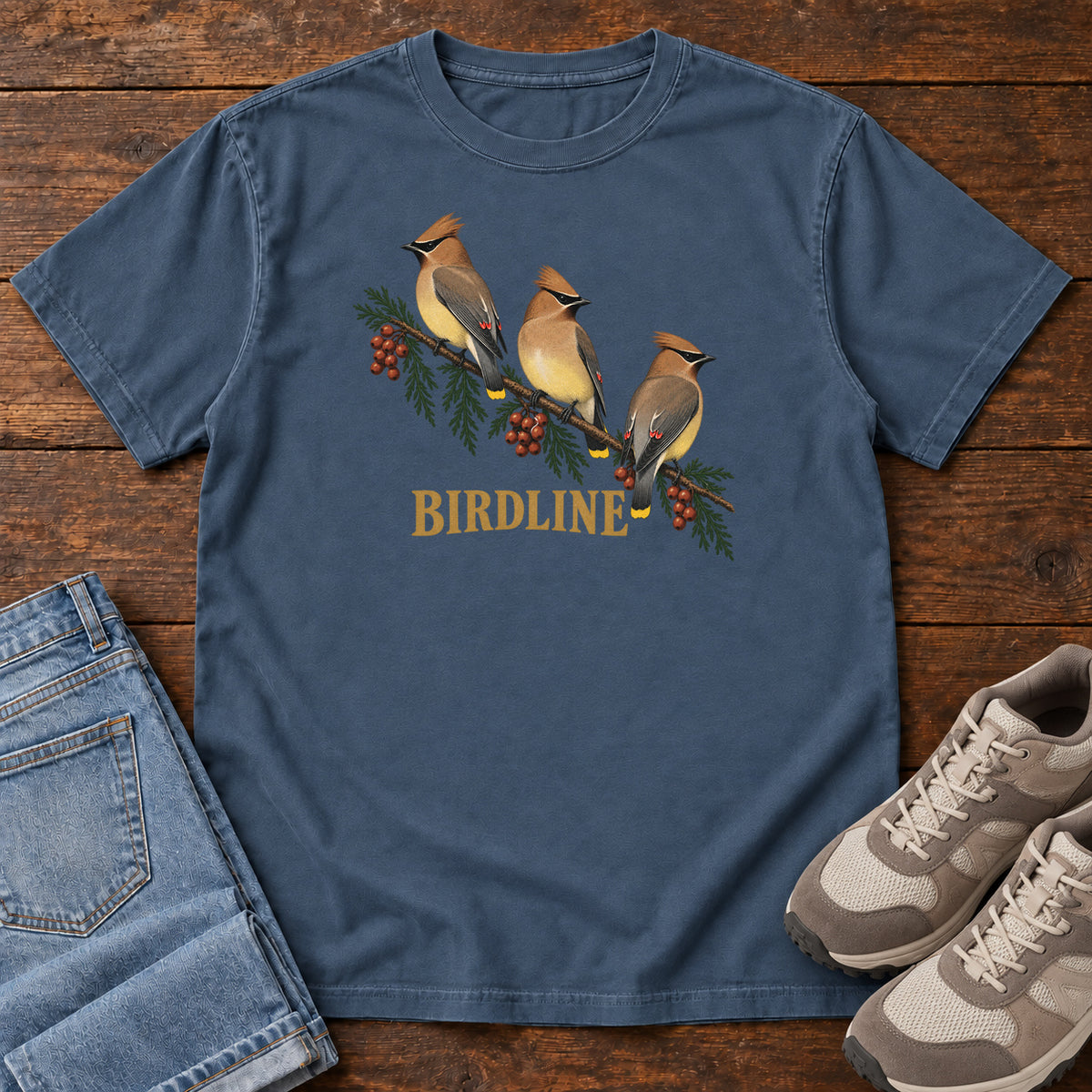 Midnight Cedar Waxwings Birdline T-Shirt with three crested birds on a berry-laden evergreen branch; front flat lay on wood.