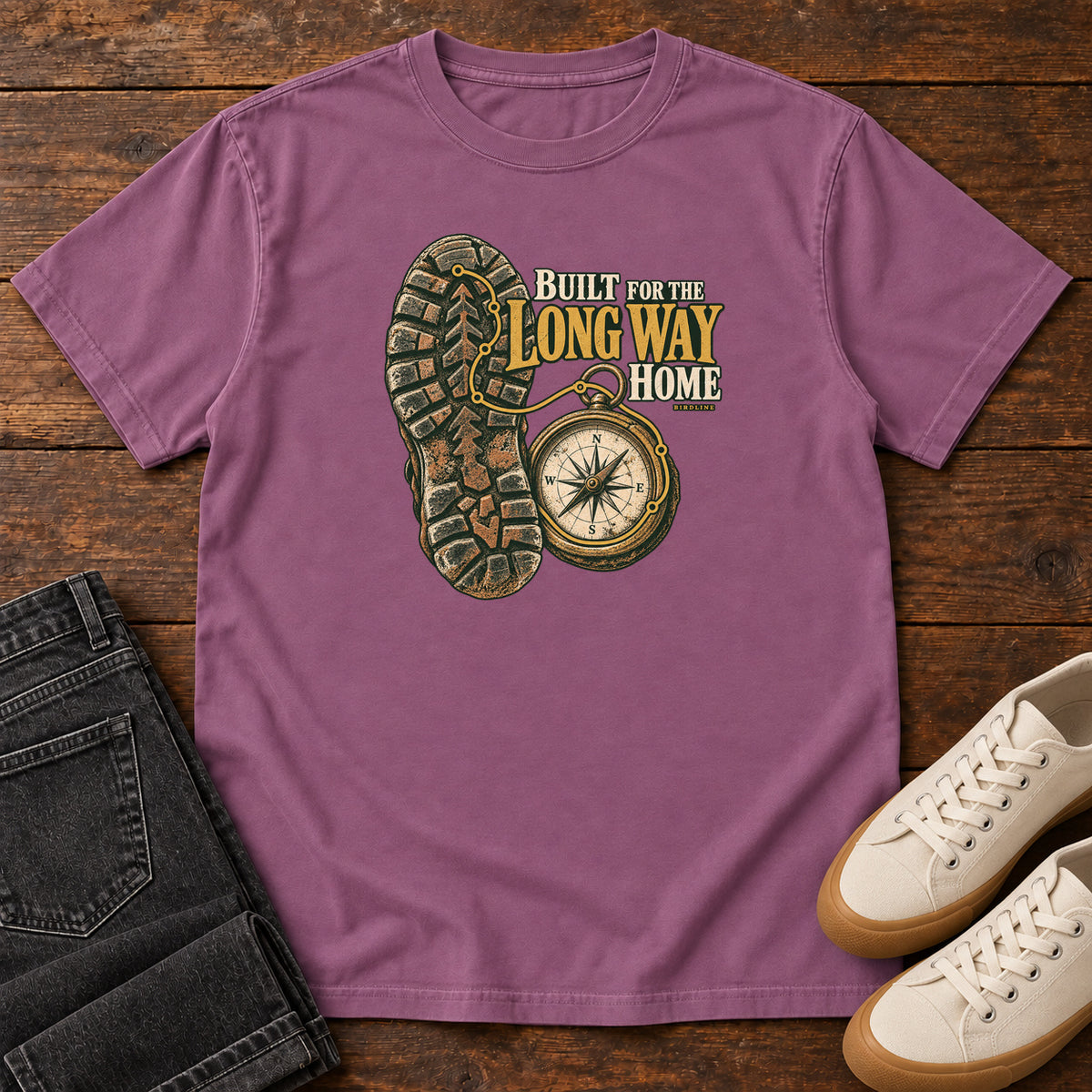 Built for the Long Way Home T-Shirt in Berry, front laid flat on wood, featuring a rugged boot sole beside a round compass and cream-and-gold slogan lettering.