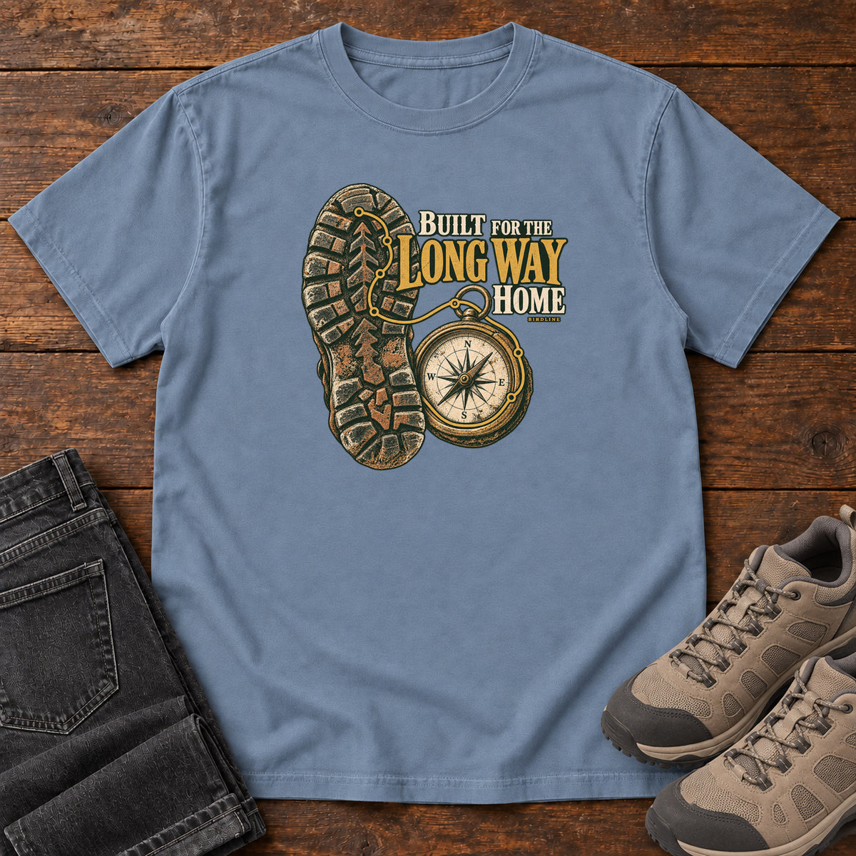 Built for the Long Way Home T-Shirt in Blue Jean, front laid flat on wood, featuring a rugged boot sole beside a round compass and cream-and-gold slogan lettering.
