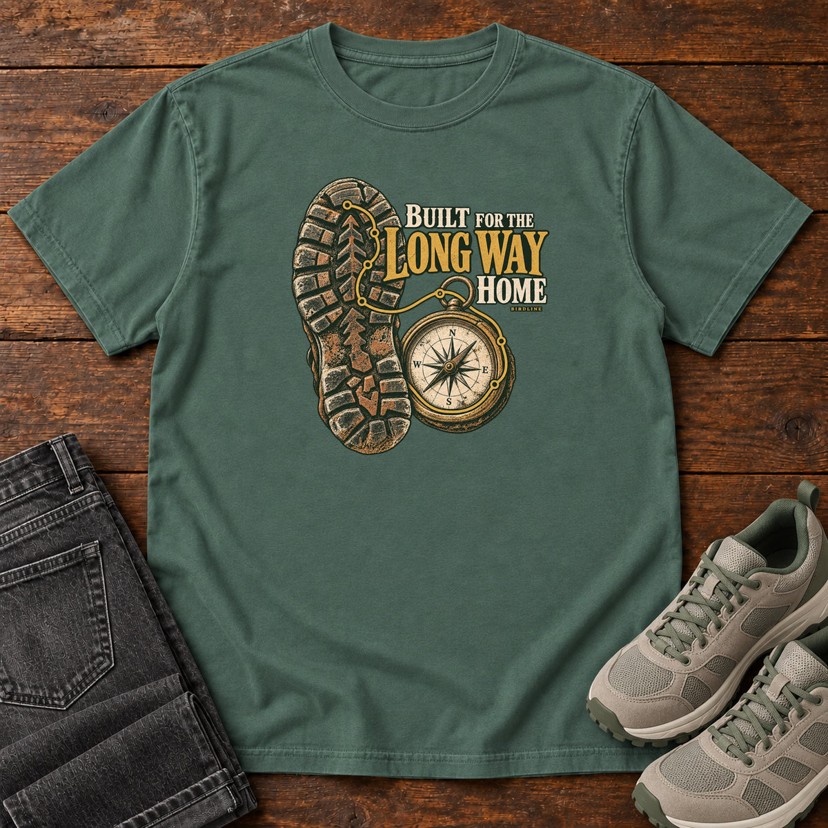 Built for the Long Way Home T-Shirt in Blue Spruce, front laid flat on wood, featuring a rugged boot sole beside a round compass and cream-and-gold slogan lettering.