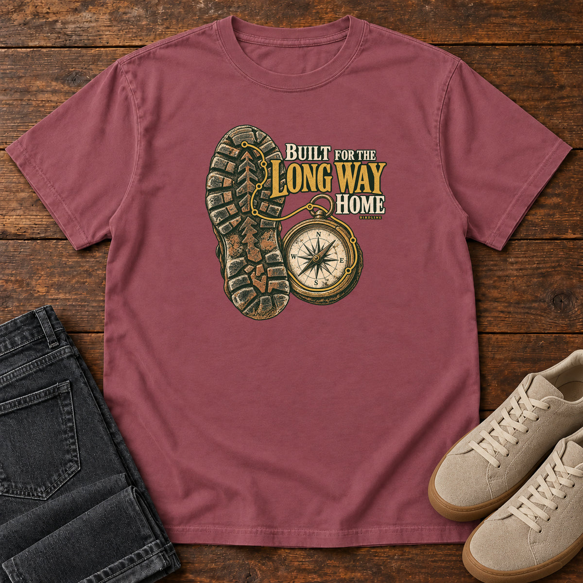 Built for the Long Way Home T-Shirt in Brick, front laid flat on wood, featuring a rugged boot sole beside a round compass and cream-and-gold slogan lettering.