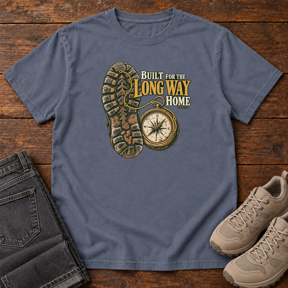 Built for the Long Way Home T-Shirt in Denim, front laid flat on wood, featuring a rugged boot sole beside a round compass and cream-and-gold slogan lettering.