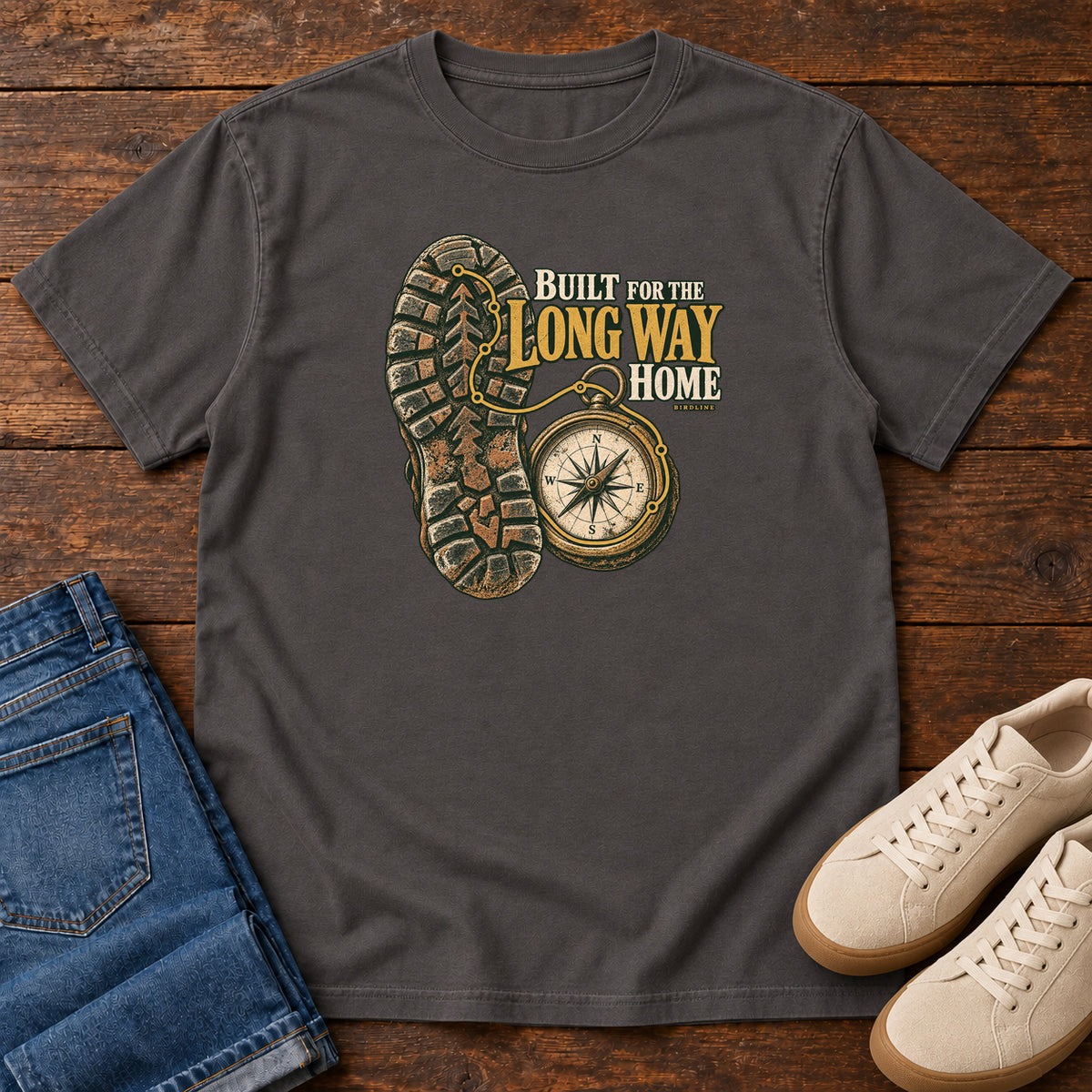 Built for the Long Way Home T-Shirt in Graphite, front laid flat on wood, featuring a rugged boot sole beside a round compass and cream-and-gold slogan lettering.
