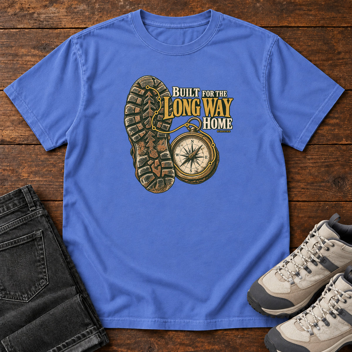 Built for the Long Way Home T-Shirt in Mystic Blue, front laid flat on wood, featuring a rugged boot sole beside a round compass and cream-and-gold slogan lettering.