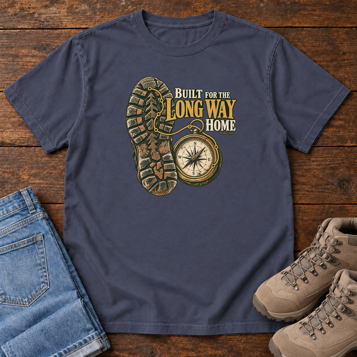 Built for the Long Way Home T-Shirt in Navy, front laid flat on wood, featuring a rugged boot sole beside a round compass and cream-and-gold slogan lettering.