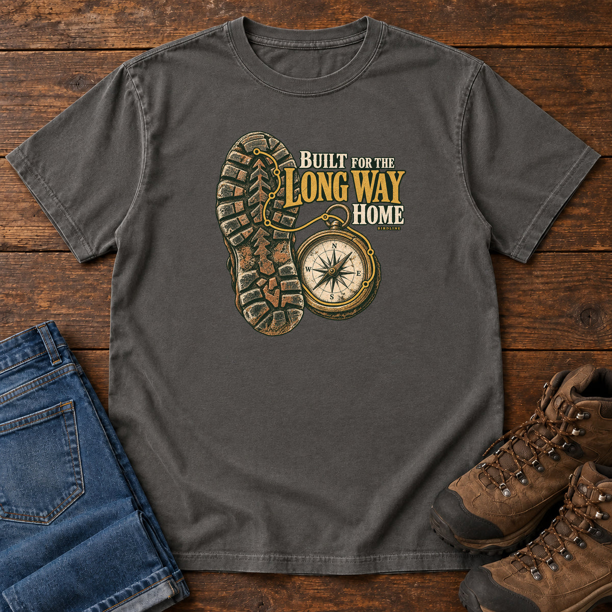 Built for the Long Way Home T-Shirt in Pepper, front laid flat on wood, featuring a rugged boot sole beside a round compass and cream-and-gold slogan lettering.