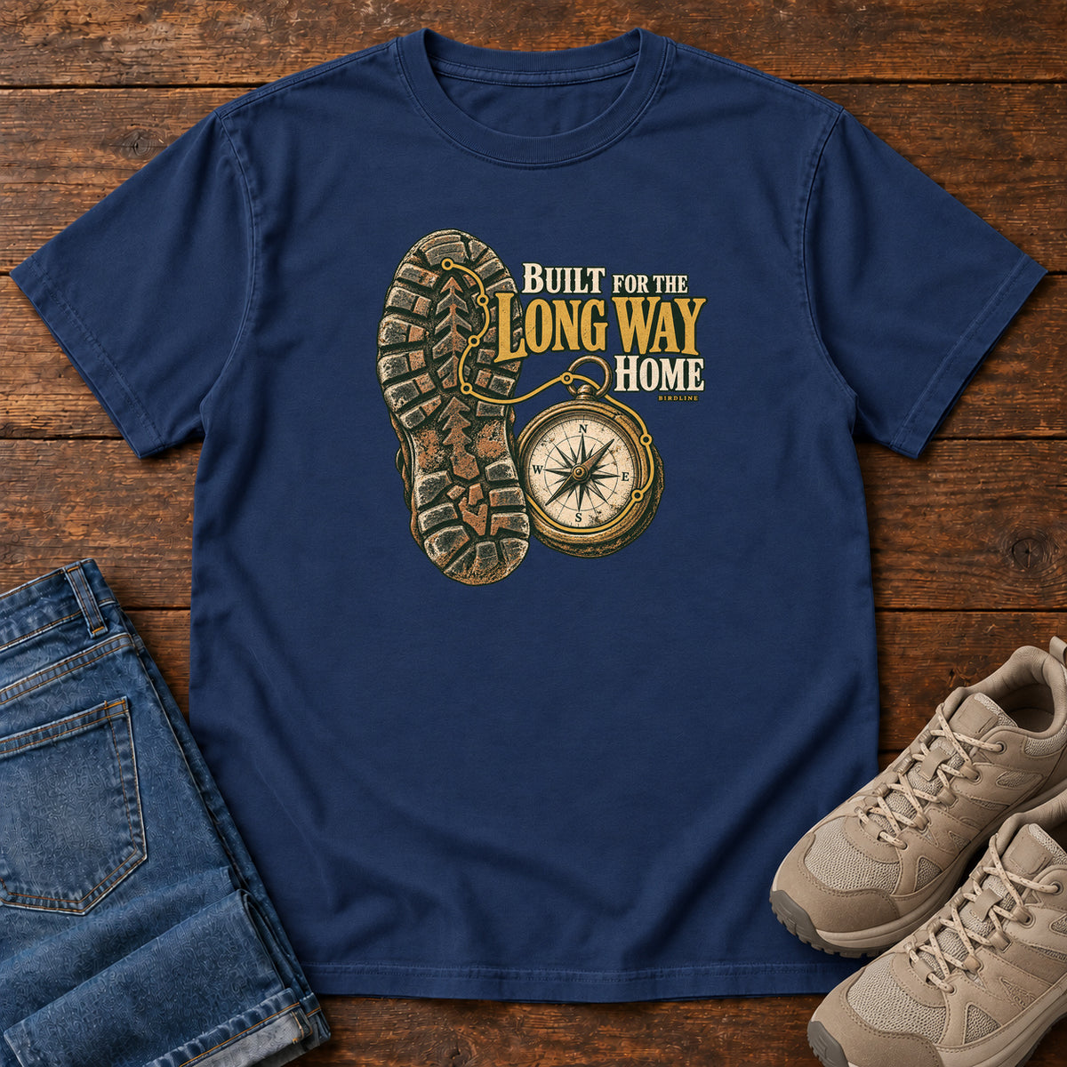 Built for the Long Way Home T-Shirt in True Navy, front laid flat on wood, featuring a rugged boot sole beside a round compass and cream-and-gold slogan lettering.