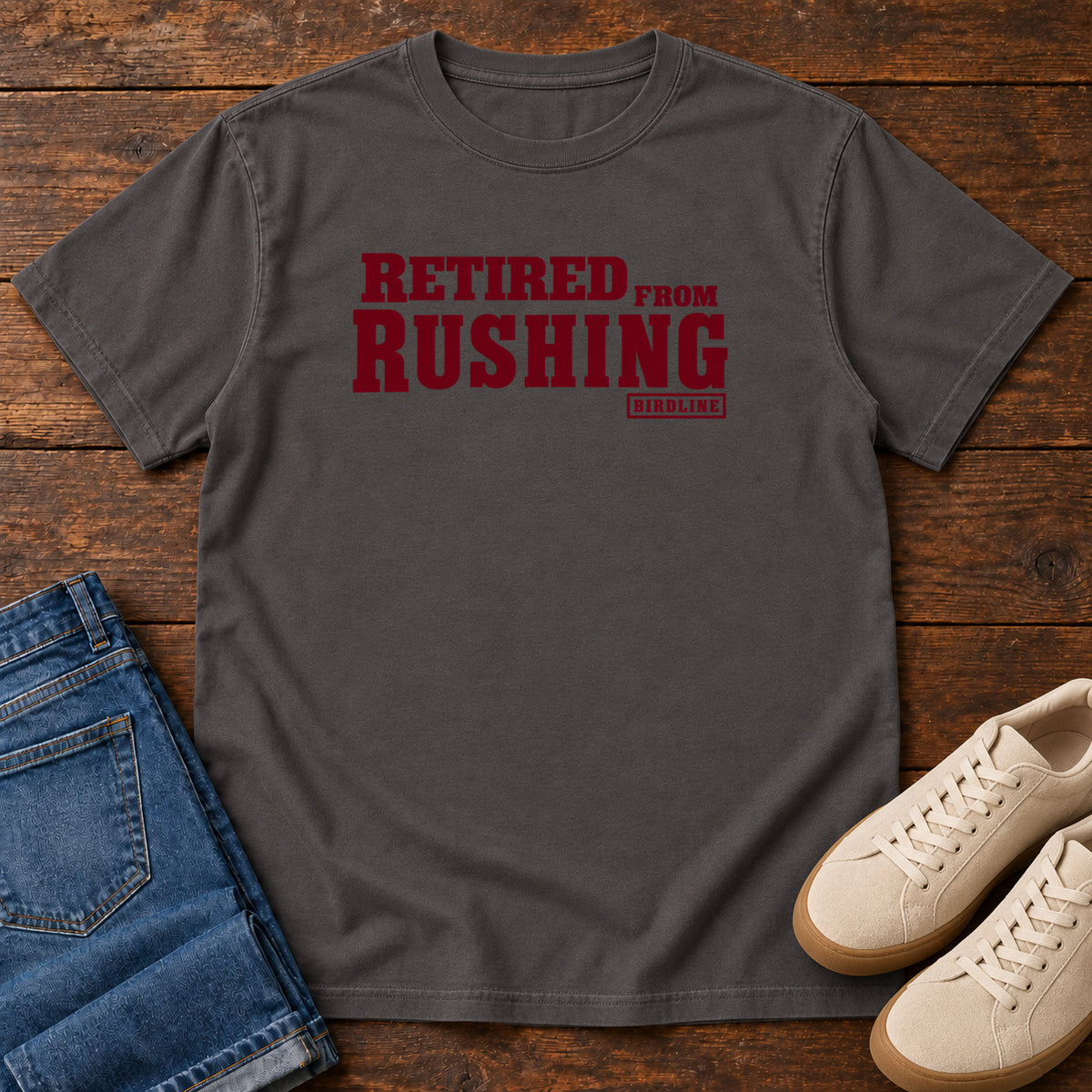 Graphite Retired From Rushing T-Shirt with burgundy Retired From Rushing lettering; front flat lay on wood.