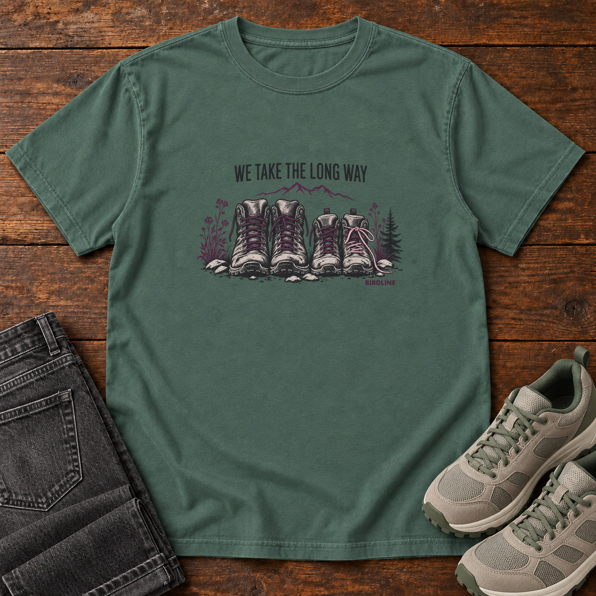 We Take the Long Way T-Shirt in Blue Spruce, front laid flat on wood, featuring two pairs of hiking boots with stones, foliage and a mountain outline beneath slogan lettering.