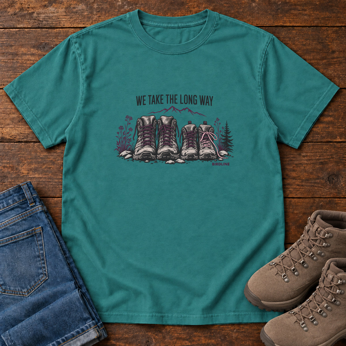 We Take the Long Way T-Shirt in Emerald, front laid flat on wood, featuring two pairs of hiking boots with stones, foliage and a mountain outline beneath slogan lettering.