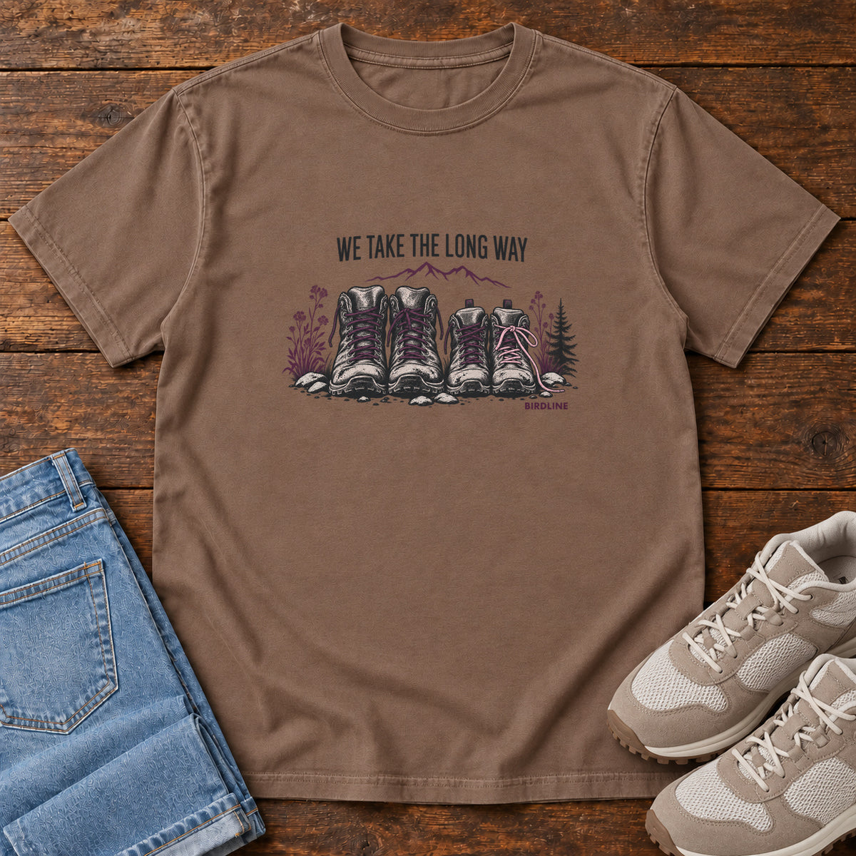 We Take the Long Way T-Shirt in Espresso, front laid flat on wood, featuring two pairs of hiking boots with stones, foliage and a mountain outline beneath slogan lettering.