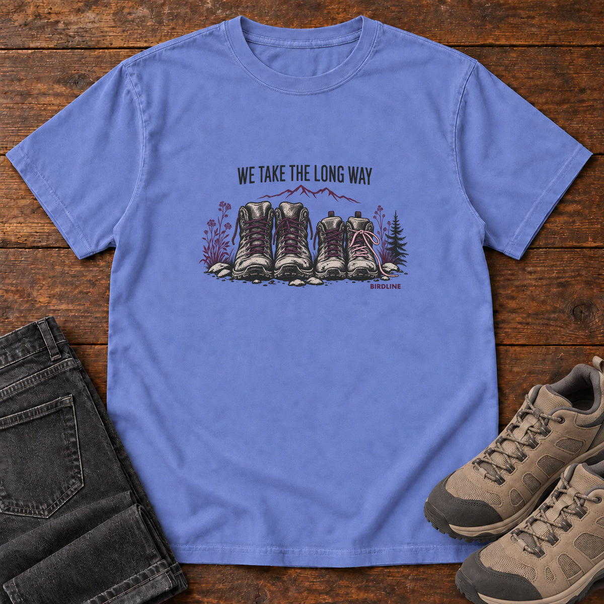 We Take the Long Way T-Shirt in Flo Blue, front laid flat on wood, featuring two pairs of hiking boots with stones, foliage and a mountain outline beneath slogan lettering.
