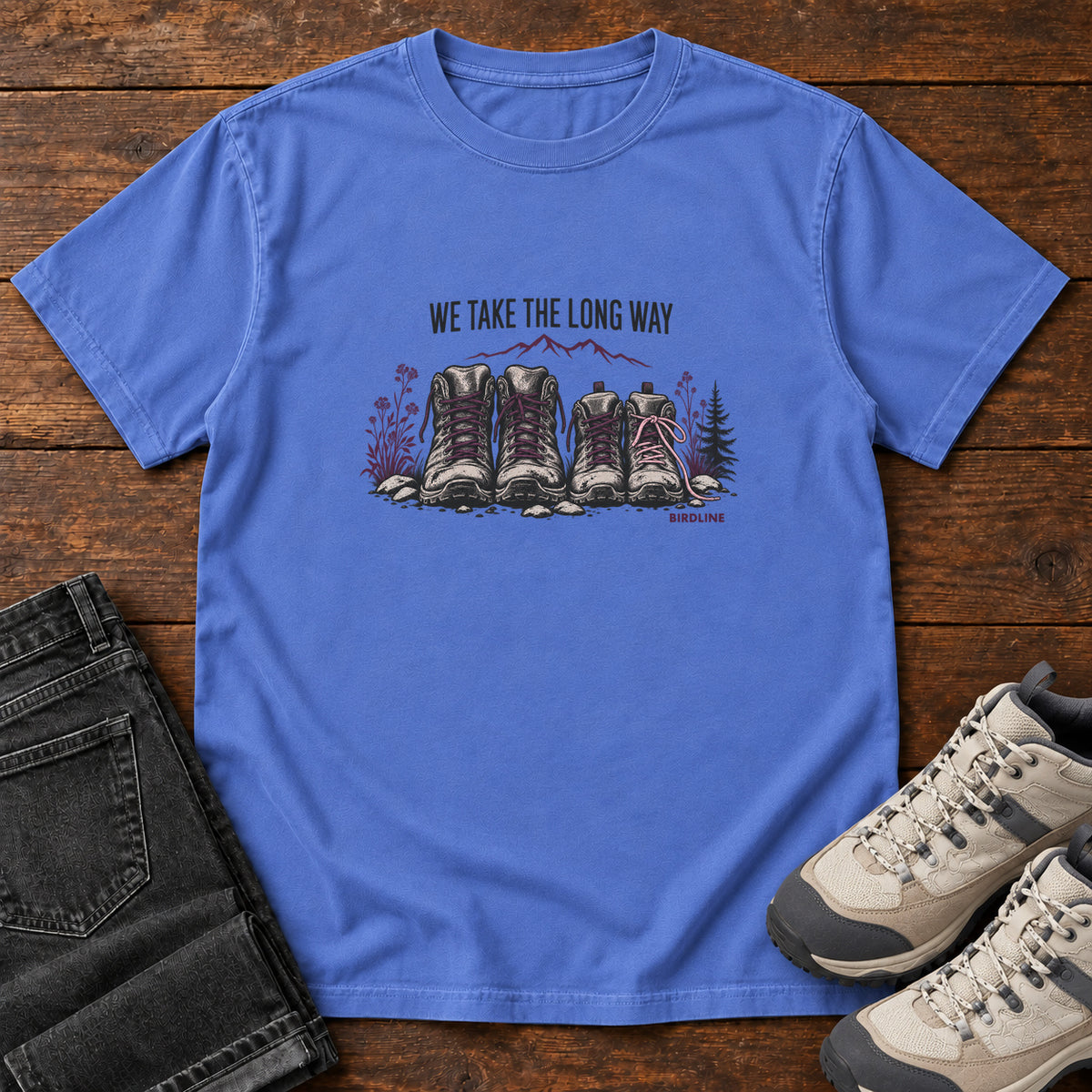 We Take the Long Way T-Shirt in Mystic Blue, front laid flat on wood, featuring two pairs of hiking boots with stones, foliage and a mountain outline beneath slogan lettering.