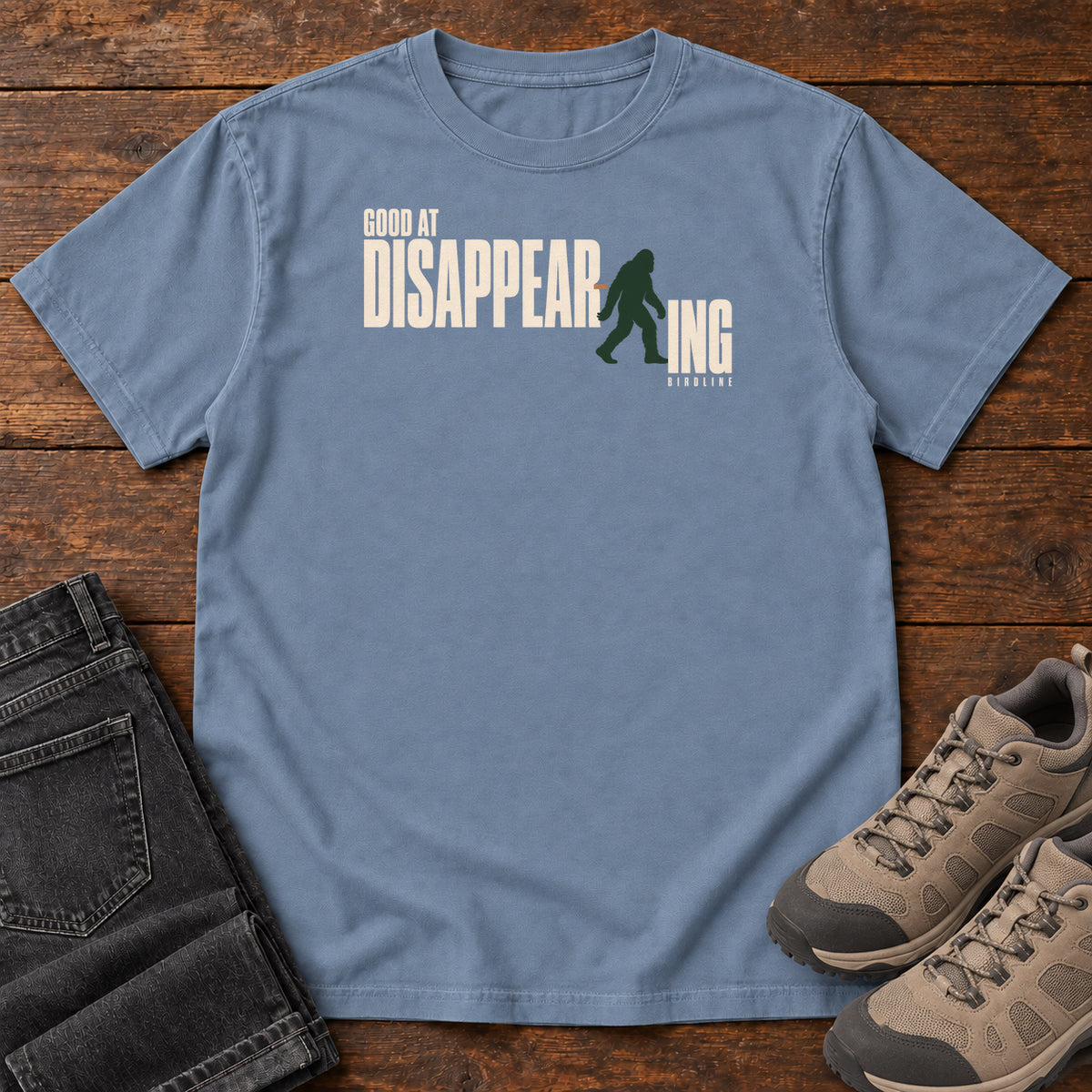 Blue Jean Good at Disappearing T-Shirt with cream lettering interrupted by a green walking Bigfoot silhouette; front flat lay on wood.