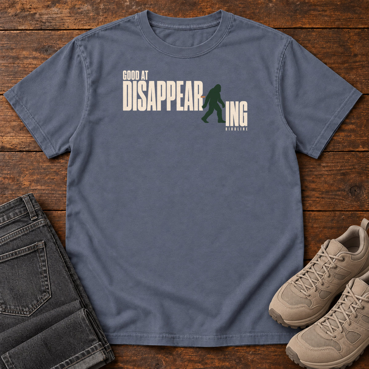 Denim Good at Disappearing T-Shirt with cream lettering interrupted by a green walking Bigfoot silhouette; front flat lay on wood.