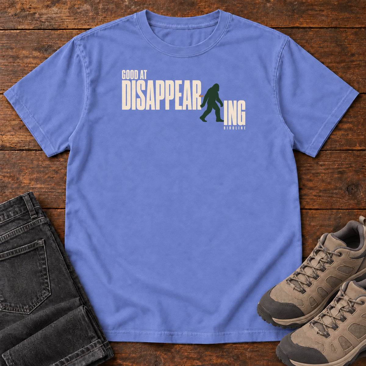 Flo Blue Good at Disappearing T-Shirt with cream lettering interrupted by a green walking Bigfoot silhouette; front flat lay on wood.