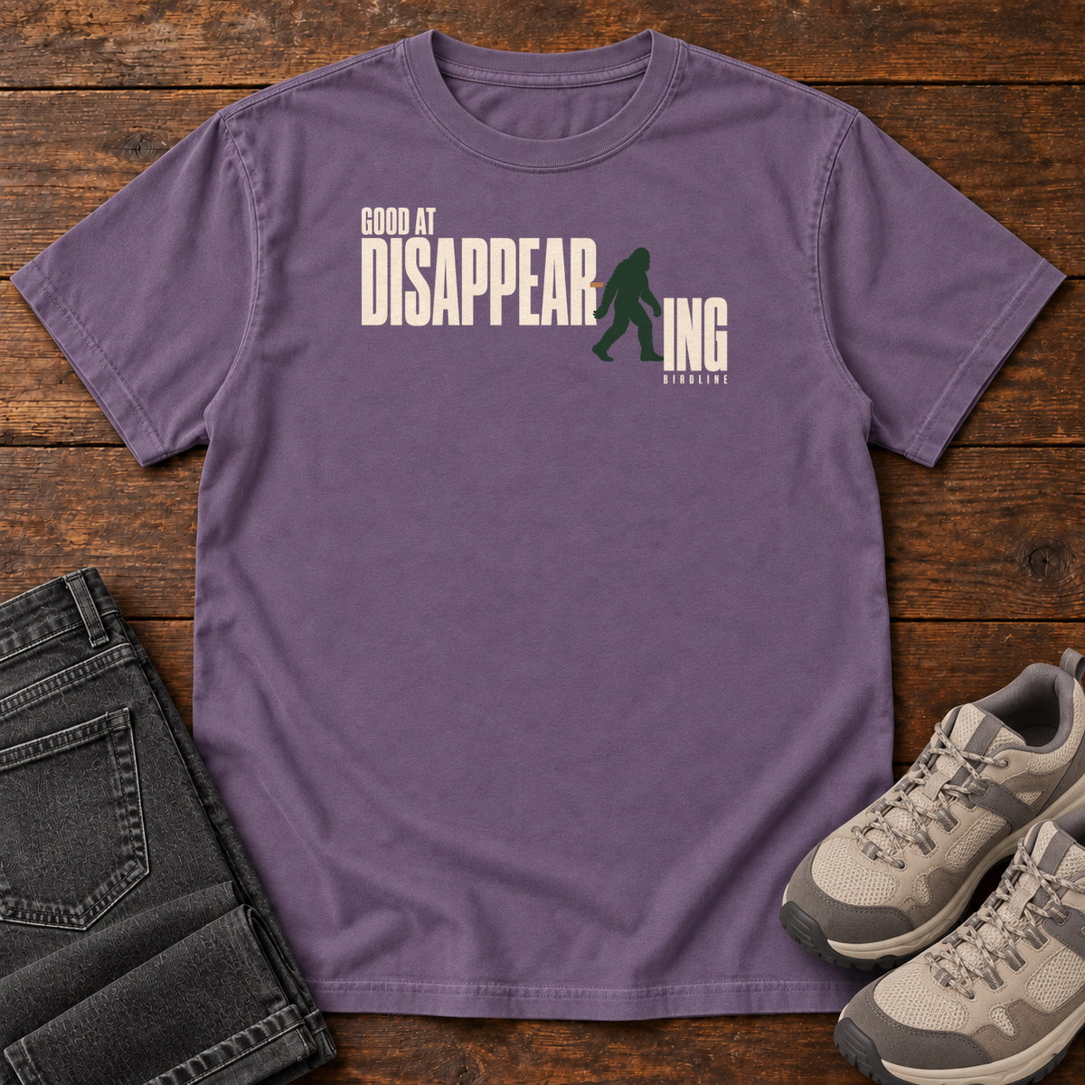 Grape Good at Disappearing T-Shirt with cream lettering interrupted by a green walking Bigfoot silhouette; front flat lay on wood.