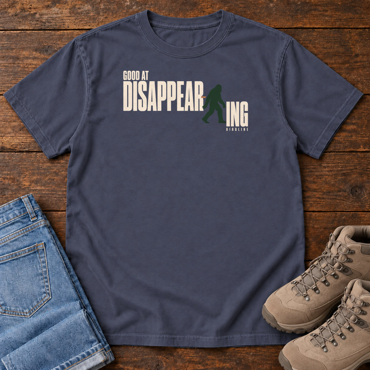 Navy Good at Disappearing T-Shirt with cream lettering interrupted by a green walking Bigfoot silhouette; front flat lay on wood.
