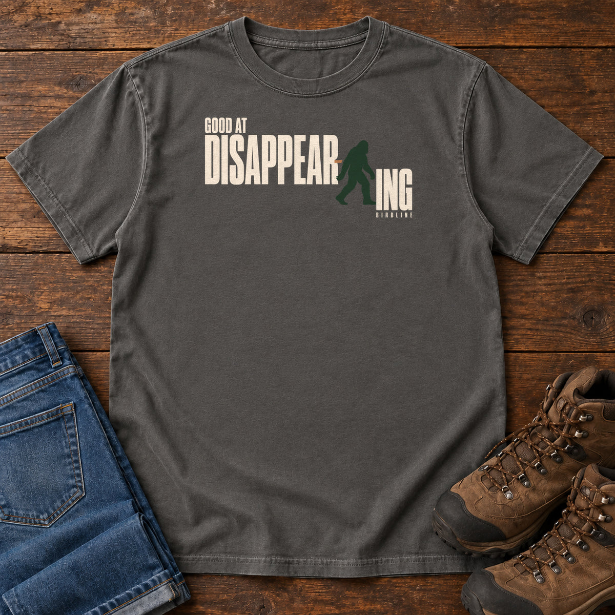 Pepper Good at Disappearing T-Shirt with cream lettering interrupted by a green walking Bigfoot silhouette; front flat lay on wood.