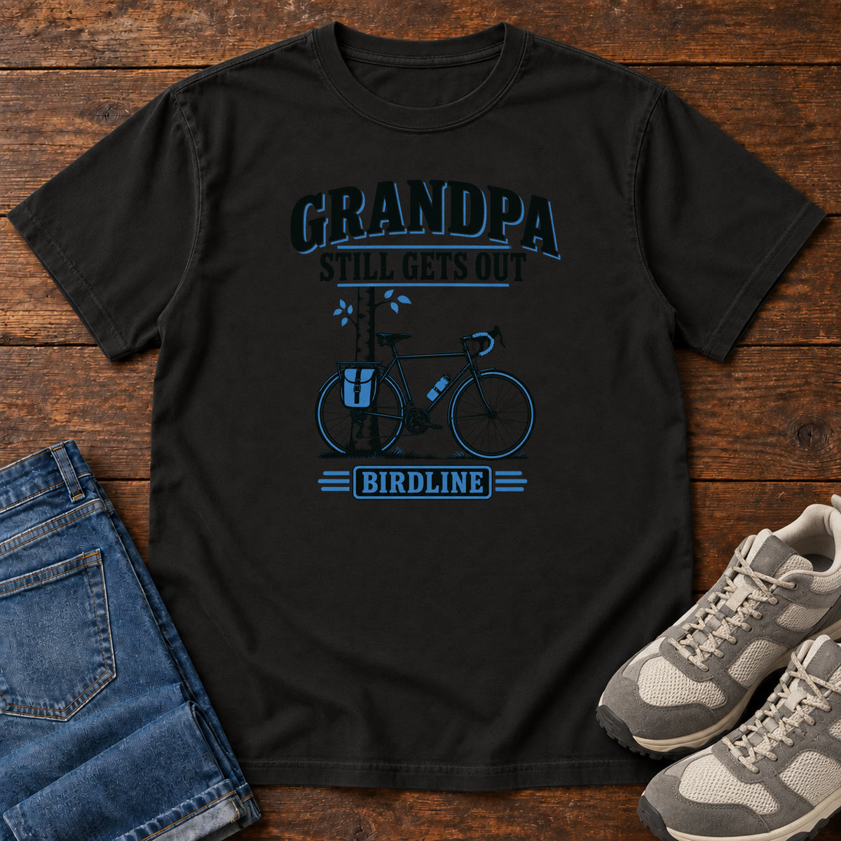 Black Grandpa Still Gets Out T-Shirt with a bicycle beside a slender tree and blue-accented lettering, front flat lay on wood.