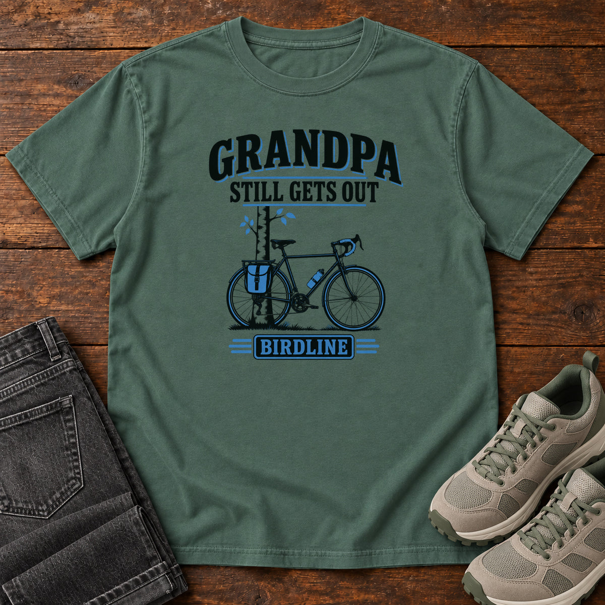 Blue Spruce Grandpa Still Gets Out T-Shirt with a bicycle beside a slender tree and blue-accented lettering, front flat lay on wood.