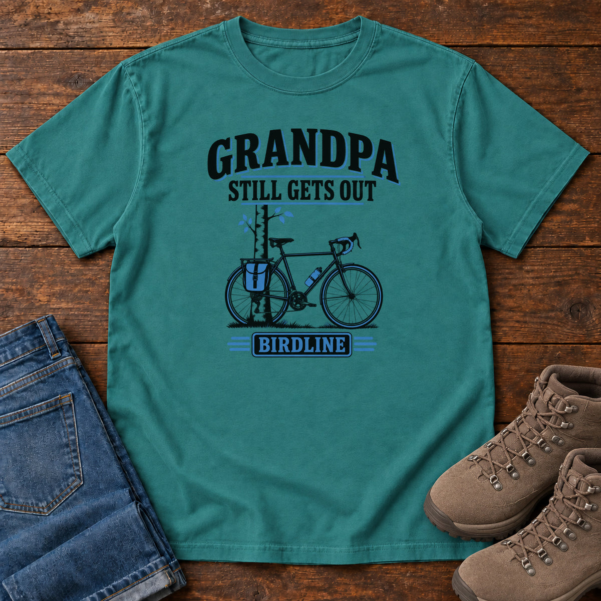Emerald Grandpa Still Gets Out T-Shirt with a bicycle beside a slender tree and blue-accented lettering, front flat lay on wood.