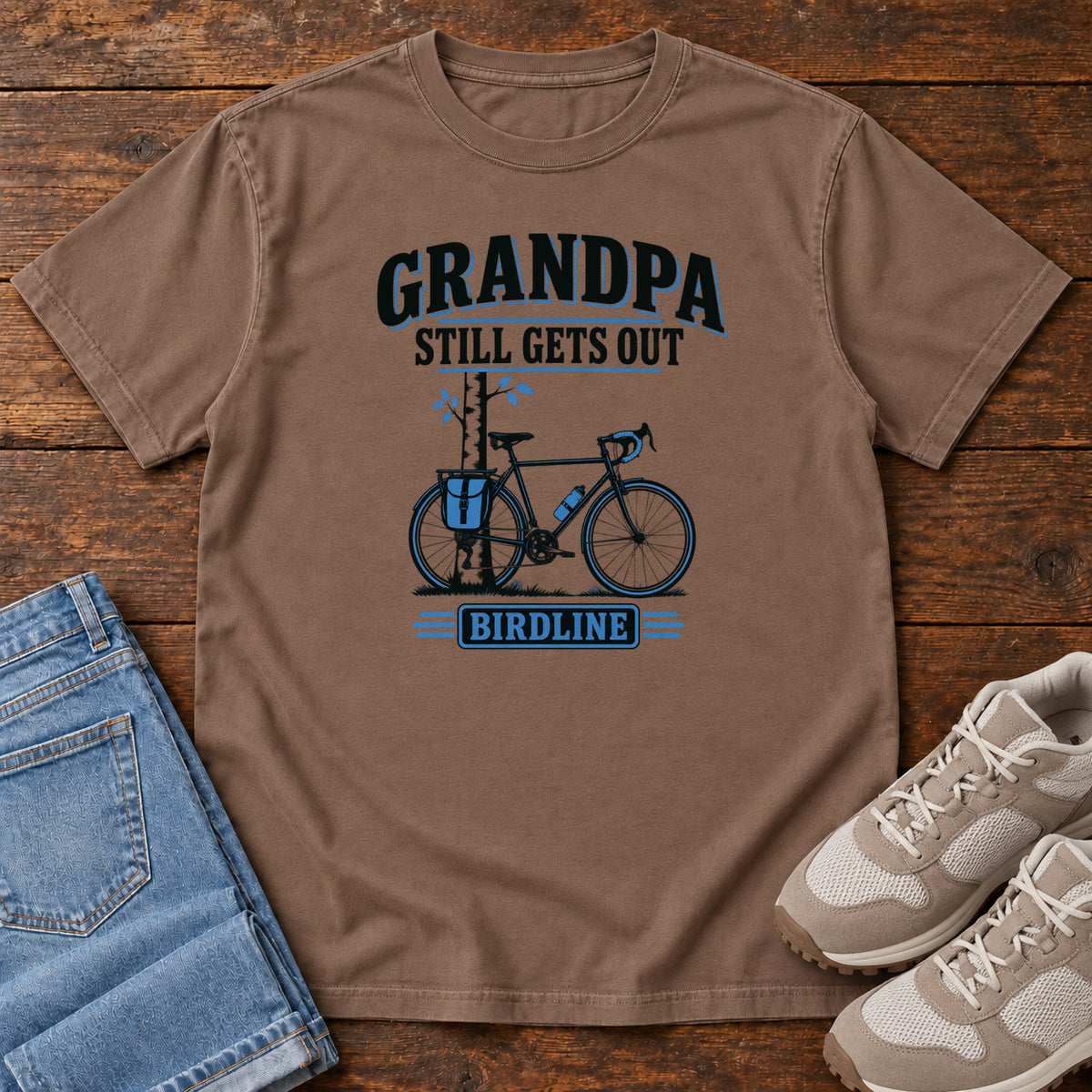 Espresso Grandpa Still Gets Out T-Shirt with a bicycle beside a slender tree and blue-accented lettering, front flat lay on wood.