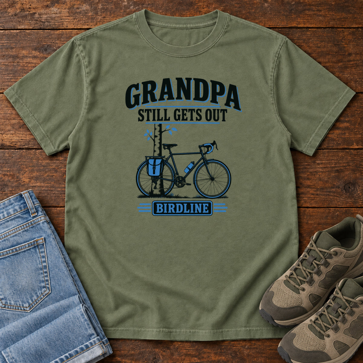Moss Grandpa Still Gets Out T-Shirt with a bicycle beside a slender tree and blue-accented lettering, front flat lay on wood.