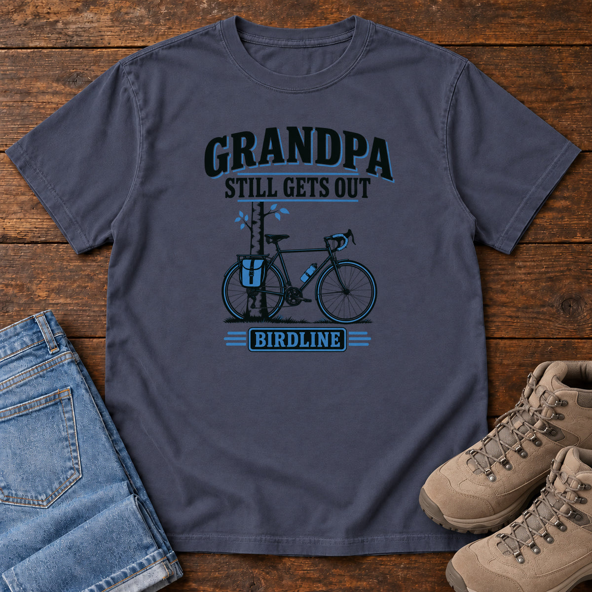 Navy Grandpa Still Gets Out T-Shirt with a bicycle beside a slender tree and blue-accented lettering, front flat lay on wood.