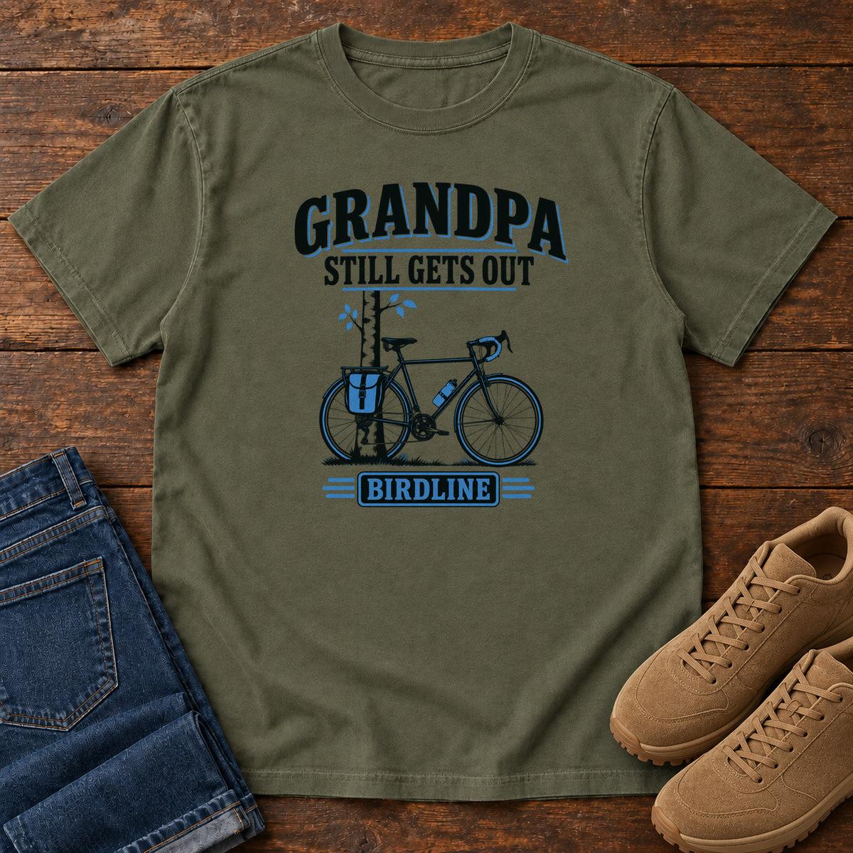 Sage Grandpa Still Gets Out T-Shirt with a bicycle beside a slender tree and blue-accented lettering, front flat lay on wood.