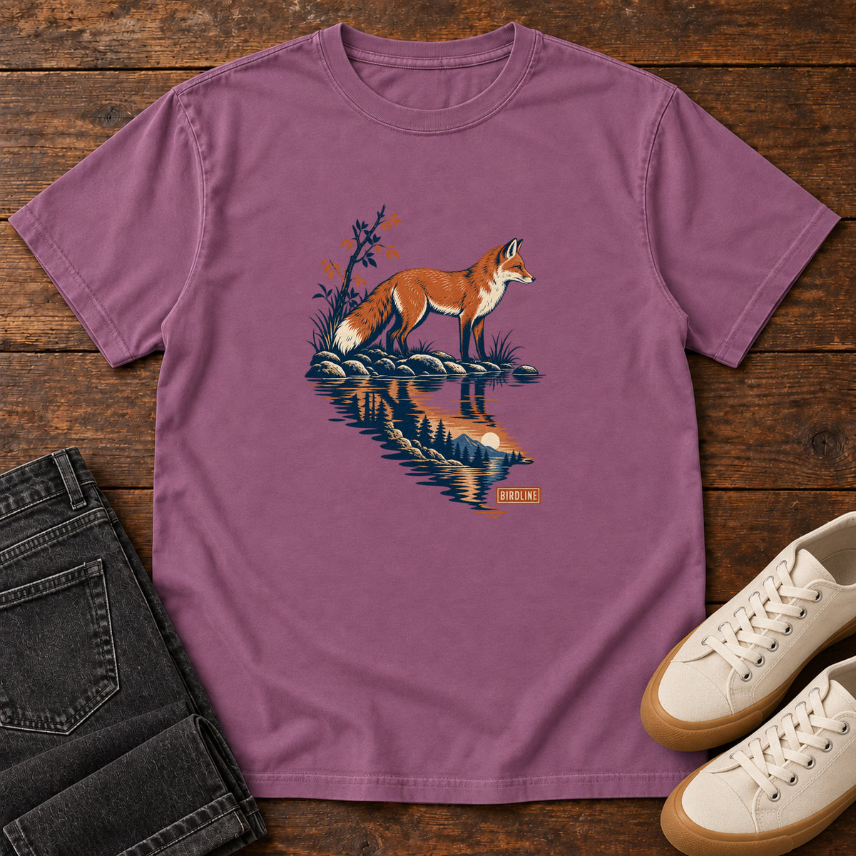 Berry Fox Reflection T-Shirt with an orange fox beside water reflecting pine trees, front flat lay on wood.