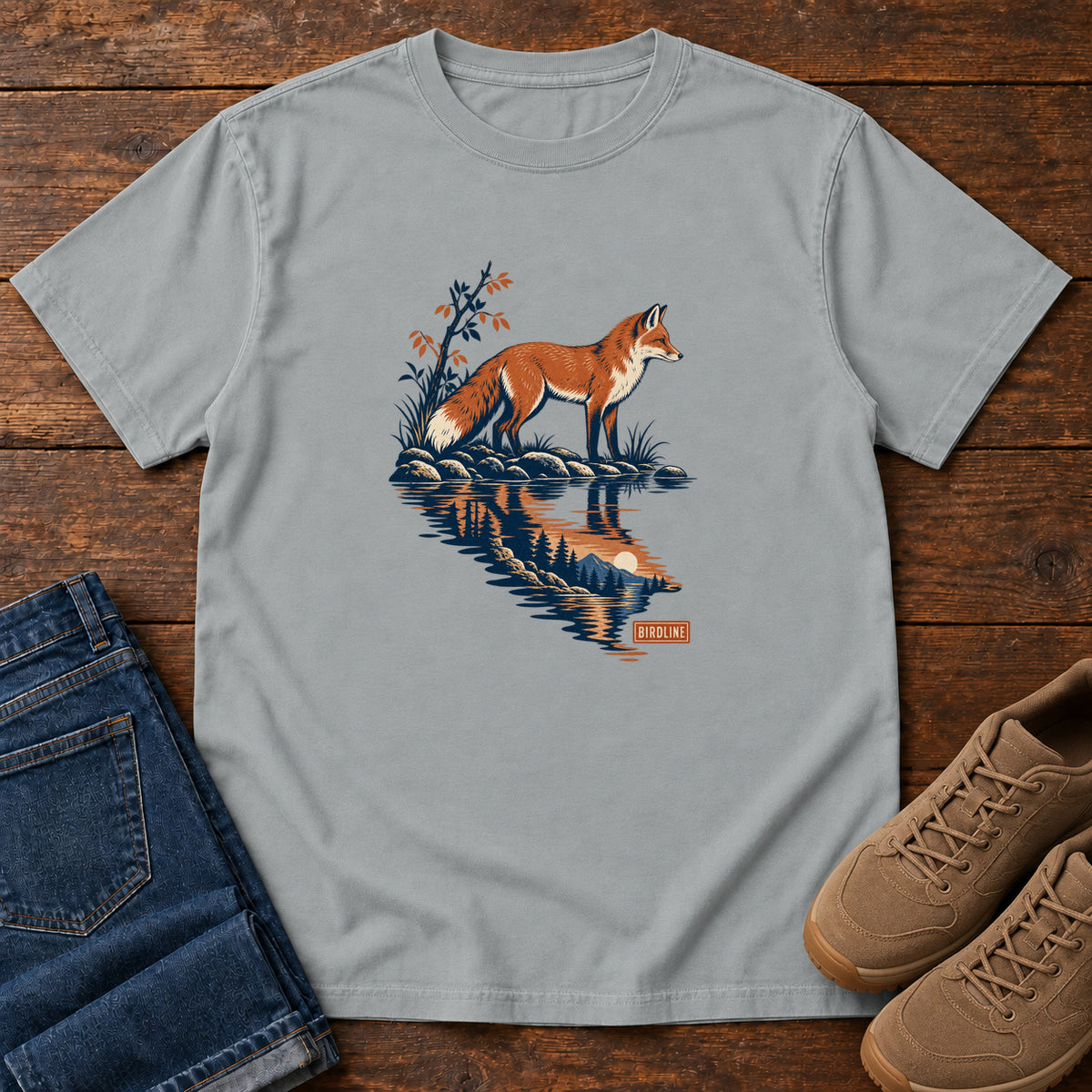 Granite Fox Reflection T-Shirt with an orange fox beside water reflecting pine trees, front flat lay on wood.