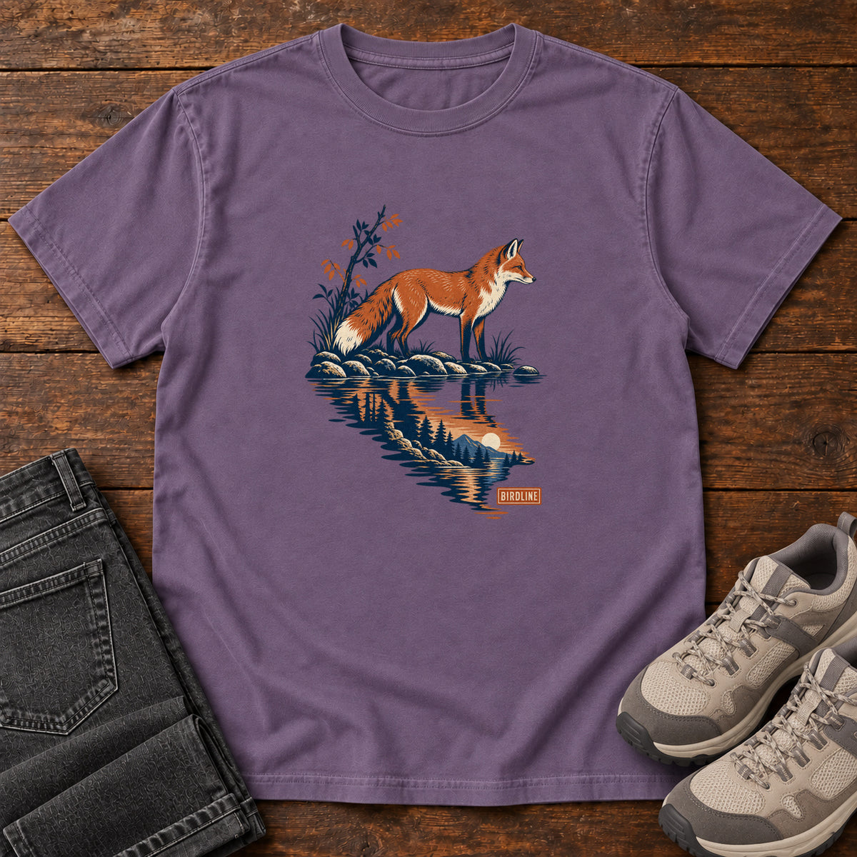 Grape Fox Reflection T-Shirt with an orange fox beside water reflecting pine trees, front flat lay on wood.