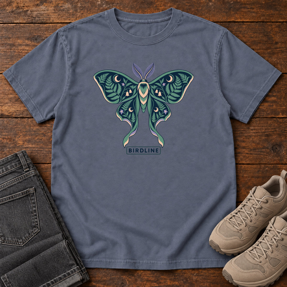 Botanical Moth T-Shirt in Denim, front laid flat on wood, featuring a teal moth with patterned wings, leaves and crescent moons.