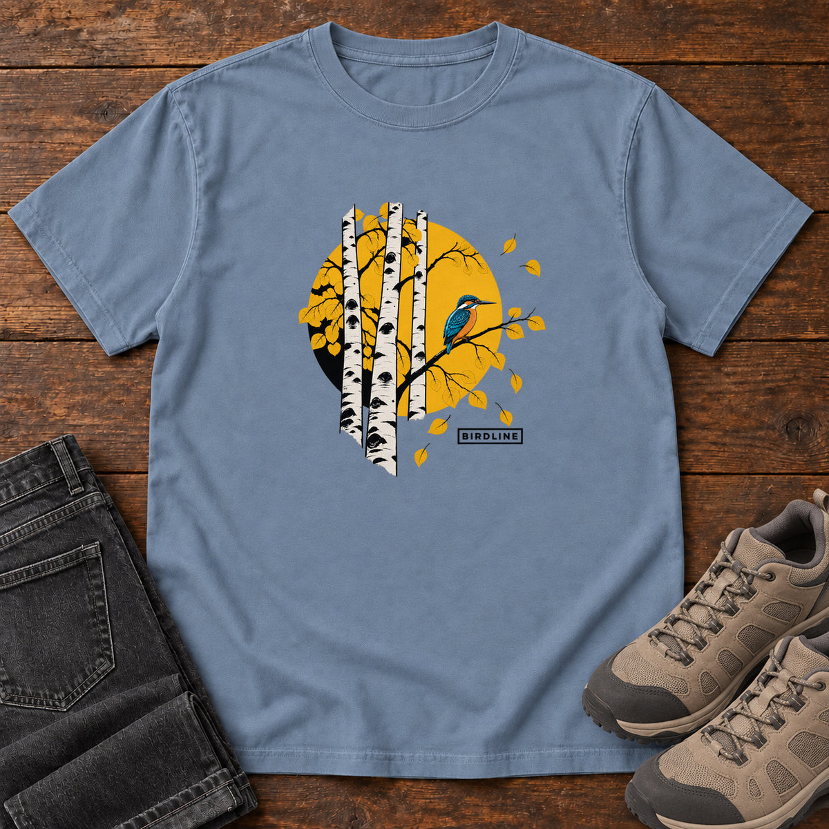 Blue Jean Aspens Kingfisher T-Shirt with a blue bird perched among white tree trunks against a golden circle, front flat lay on wood.