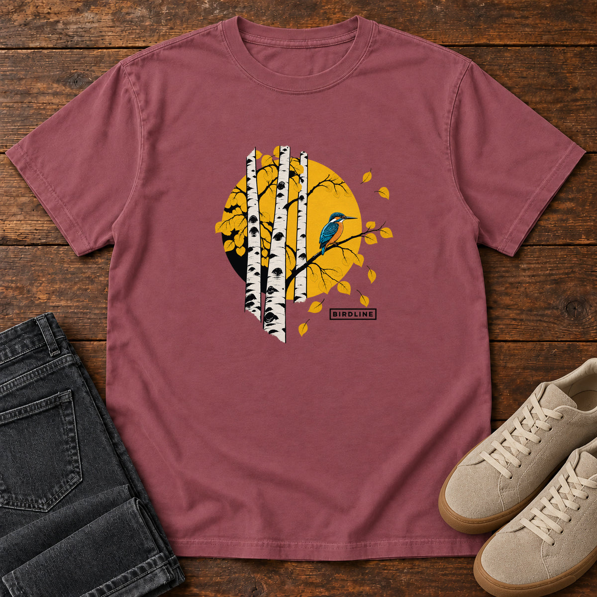 Brick Aspens Kingfisher T-Shirt with a blue bird perched among white tree trunks against a golden circle, front flat lay on wood.