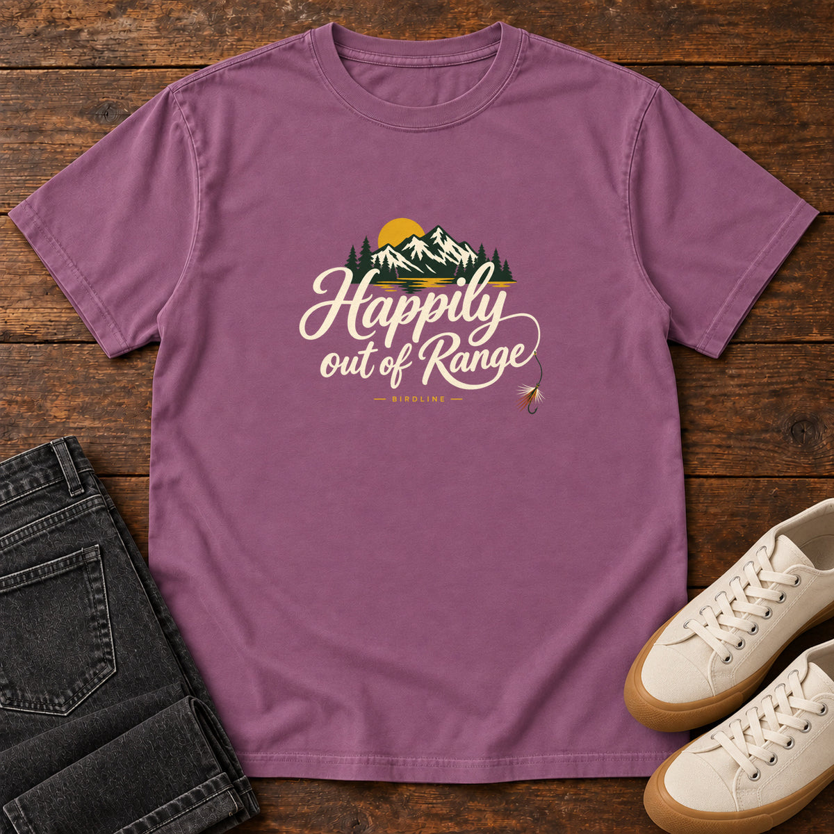 Berry Happily out of Range T-Shirt with mountains, evergreens and a golden sun above cream script lettering; front flat lay on wood.