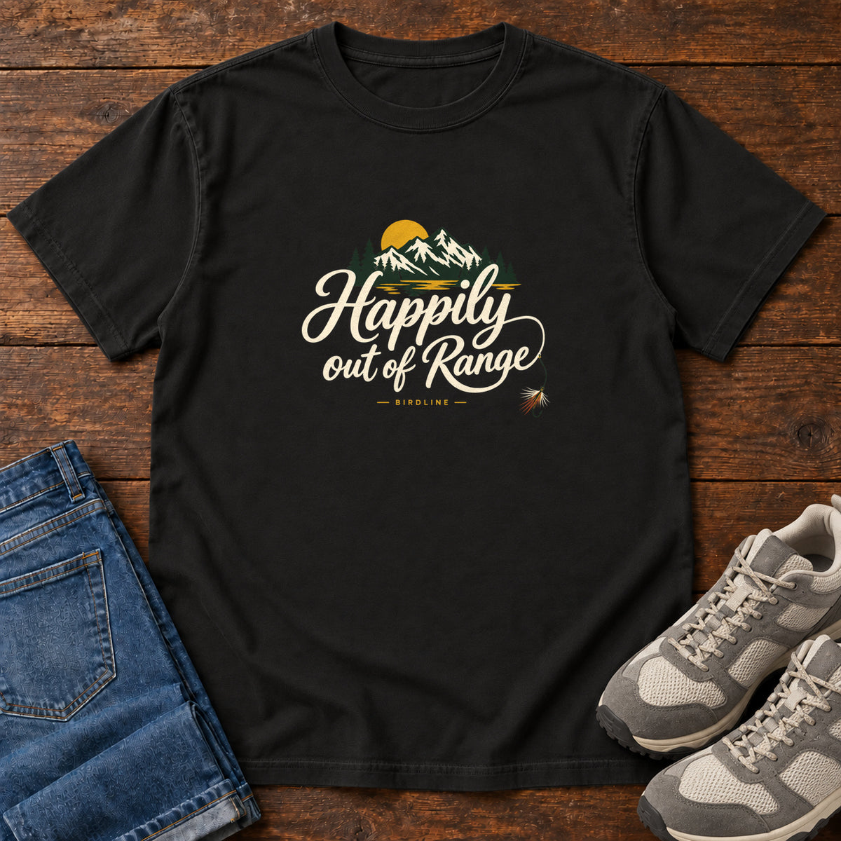 Black Happily out of Range T-Shirt with mountains, evergreens and a golden sun above cream script lettering; front flat lay on wood.