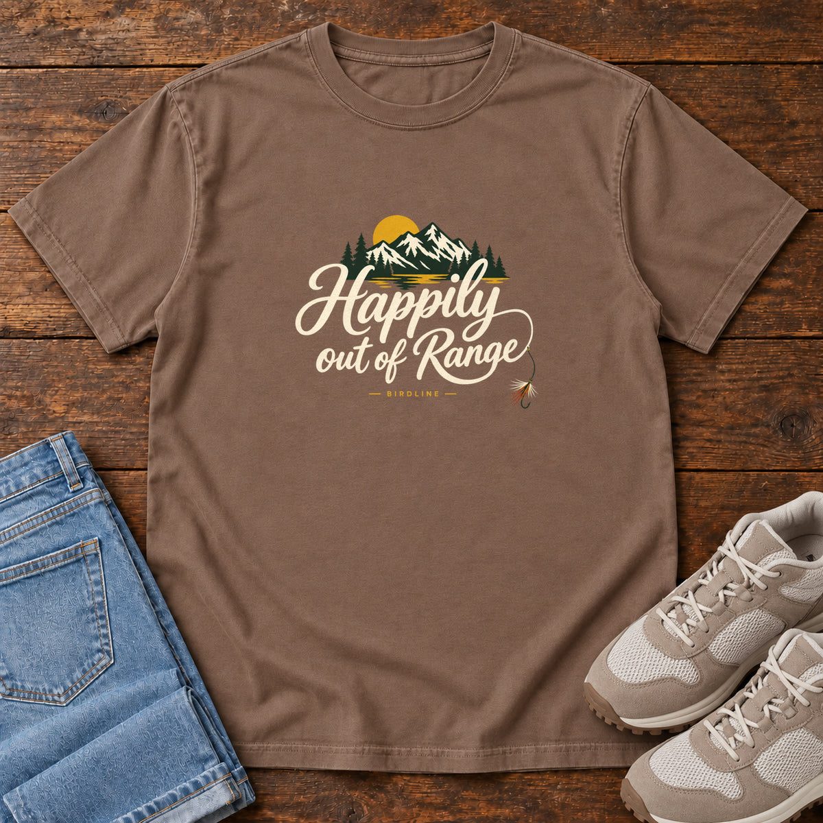 Espresso Happily out of Range T-Shirt with mountains, evergreens and a golden sun above cream script lettering; front flat lay on wood.