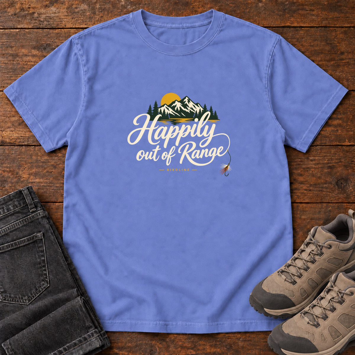 Flo Blue Happily out of Range T-Shirt with mountains, evergreens and a golden sun above cream script lettering; front flat lay on wood.