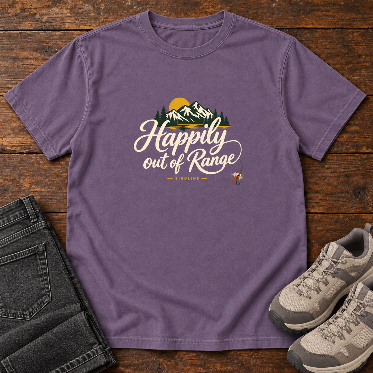 Grape Happily out of Range T-Shirt with mountains, evergreens and a golden sun above cream script lettering; front flat lay on wood.