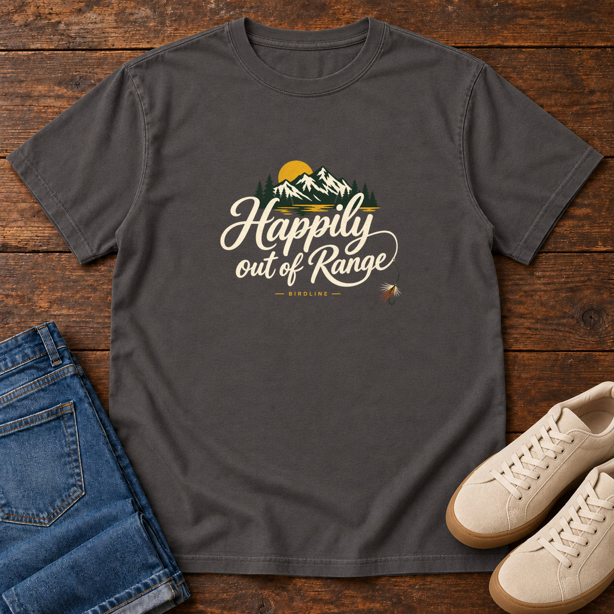 Graphite Happily out of Range T-Shirt with mountains, evergreens and a golden sun above cream script lettering; front flat lay on wood.