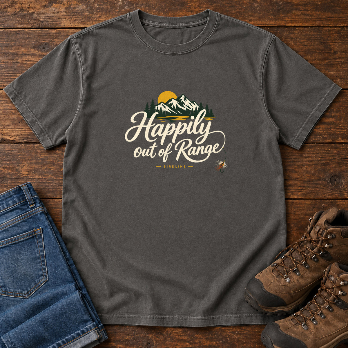 Pepper Happily out of Range T-Shirt with mountains, evergreens and a golden sun above cream script lettering; front flat lay on wood.