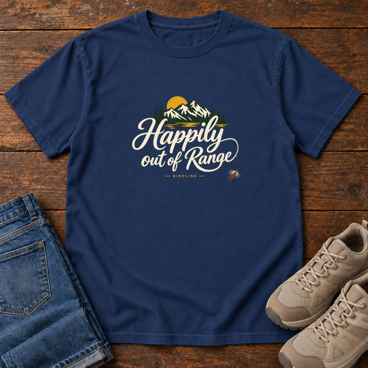 True Navy Happily out of Range T-Shirt with mountains, evergreens and a golden sun above cream script lettering; front flat lay on wood.