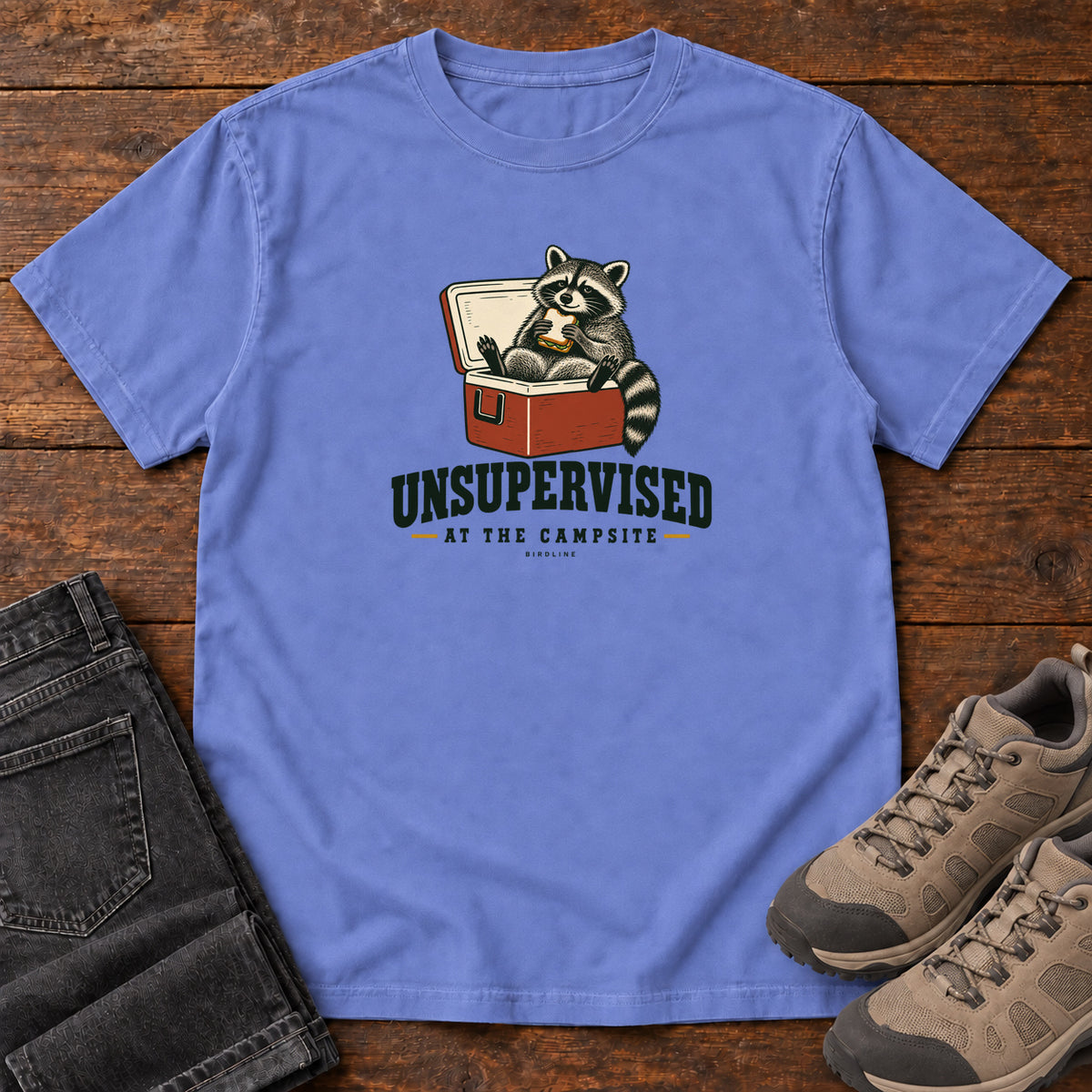 Flo Blue Unsupervised at the Campsite T-Shirt with a raccoon eating a sandwich while sitting in an open red cooler; front flat lay on wood.
