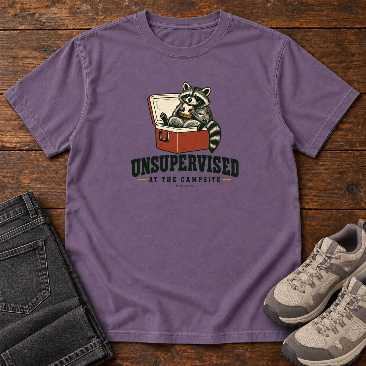 Grape Unsupervised at the Campsite T-Shirt with a raccoon eating a sandwich while sitting in an open red cooler; front flat lay on wood.