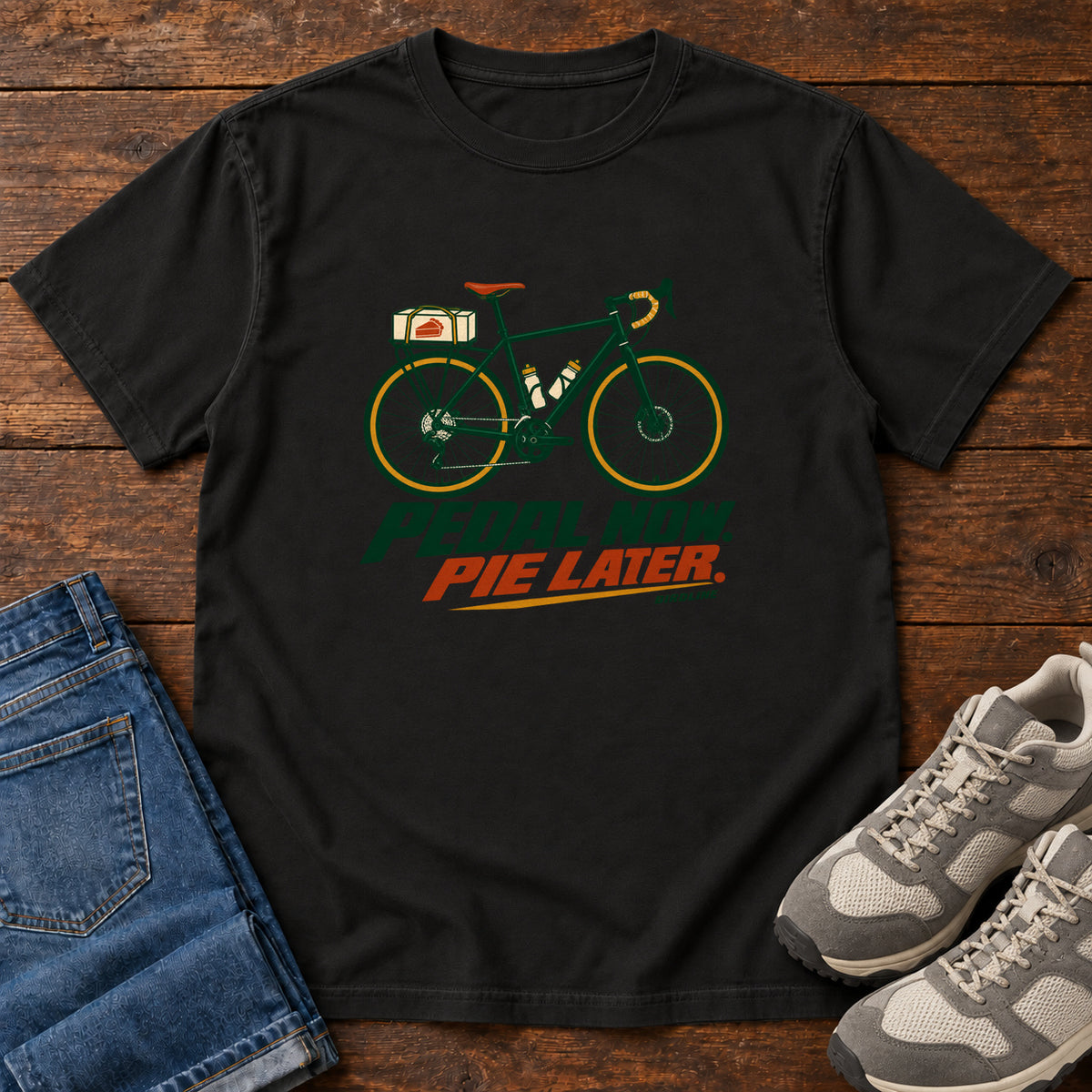 Black Pedal Now Pie Later T-Shirt with a green touring bicycle above green and orange lettering, front flat lay on wood.