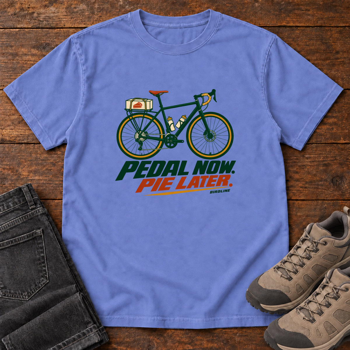 Flo Blue Pedal Now Pie Later T-Shirt with a green touring bicycle above green and orange lettering, front flat lay on wood.