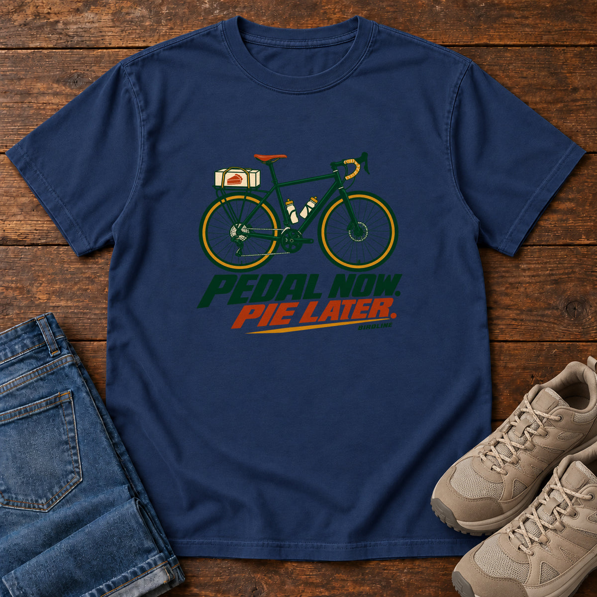 True Navy Pedal Now Pie Later T-Shirt with a green touring bicycle above green and orange lettering, front flat lay on wood.