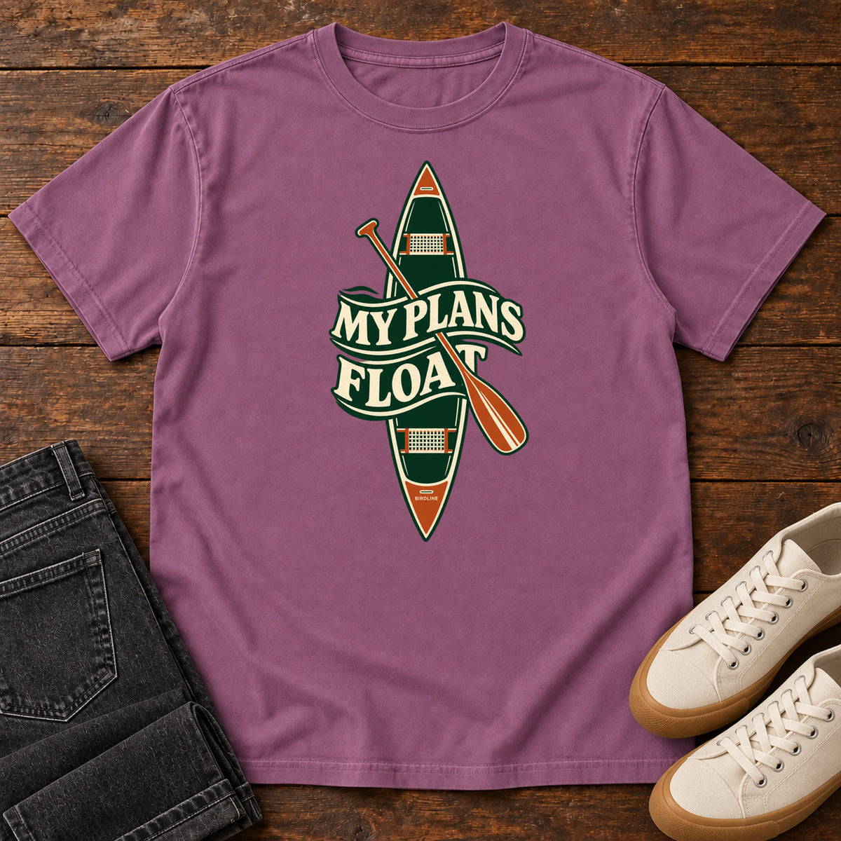 My Plans Float T-Shirt in Berry, front laid flat on wood, featuring a vertical green canoe and orange paddle behind cream slogan lettering.